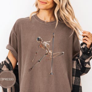 May include: Espresso-colored t-shirt featuring a skeleton pole dancing. The skeleton is depicted in a dynamic pose, holding onto a silver pole. The shirt has a relaxed fit and is made of a soft material. The word "ESPRESSO" is printed on a brown circle.