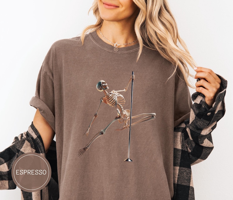 May include: Espresso-colored t-shirt featuring a skeleton pole dancing. The skeleton is depicted in a dynamic pose, holding onto a silver pole. The shirt has a relaxed fit and is made of a soft material. The word "ESPRESSO" is printed on a brown circle.