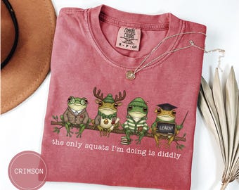 Könnte beinhalten: Karmesinrotes T-Shirt mit einer humorvollen Illustration von vier Fröschen in verschiedenen Outfits, die auf einem Ast sitzen. Der Text "the only squats I'm doing is diddly" ist darunter gedruckt. Eine goldene Halskette liegt auf dem T-Shirt.