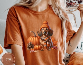 Op de afbeelding: Oranje T-shirt met een teckel die een bruine en oranje hoed en sjaal draagt, omringd door pompoenen en herfstbladeren. De tekst "sam" is op het T-shirt gedrukt.