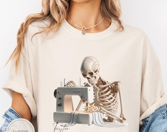 Könnte beinhalten: Elfenbeinfarbenes T-Shirt mit einem Skelett, das eine weiße Nähmaschine bedient. Das Skelett ist mit detaillierter Knochenstruktur dargestellt. Der Name "Taylor" steht in Schreibschrift darunter. Das Shirt ist ein Freizeitartikel.