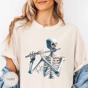 Peut inclure: T-shirt ivoire avec une illustration détaillée d'un squelette jouant de la flûte. Le squelette est rendu en blanc et bleu. Le nom "Taylor" est écrit en cursive. Le t-shirt est de couleur neutre.