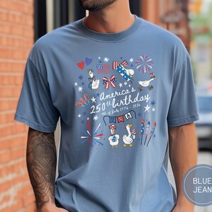 May include: Blue jean-coloured t-shirt featuring a graphic celebrating America's 250th birthday. The design includes cartoon geese, fireworks, and patriotic elements in red, white, and blue. Text reads "America's 250th birthday 4th of July 1776-2026".