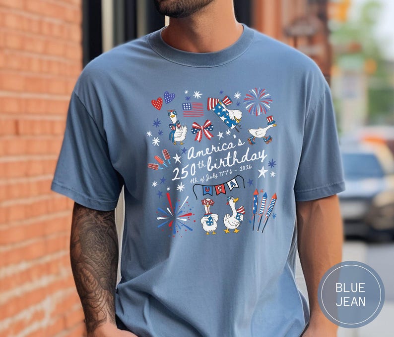 May include: Blue jean-coloured t-shirt featuring a graphic celebrating America's 250th birthday. The design includes cartoon geese, fireworks, and patriotic elements in red, white, and blue. Text reads "America's 250th birthday 4th of July 1776-2026".