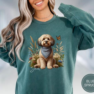 May include: Teal blue sweatshirt with a graphic of a brown and white dog wearing a blue bandana. The dog is surrounded by flowers and a butterfly. The text "Sam" is written below the dog.