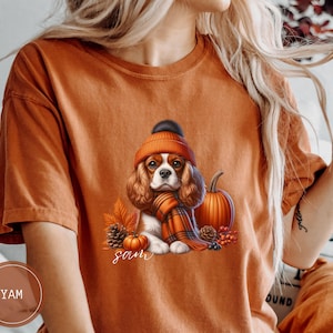 May include: Orange t-shirt with a graphic of a Cavalier King Charles Spaniel wearing a brown and white hat and a red and black scarf. The dog is sitting next to a pumpkin and pine cones. The text "Sam" is printed below the dog.