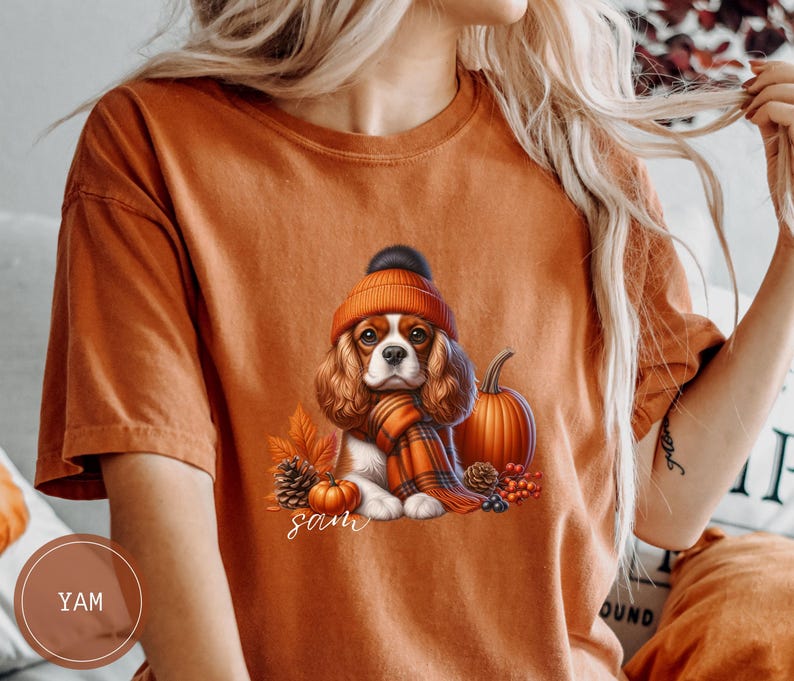 May include: Orange t-shirt with a graphic of a Cavalier King Charles Spaniel wearing a brown and white hat and a red and black scarf. The dog is sitting next to a pumpkin and pine cones. The text "Sam" is printed below the dog.