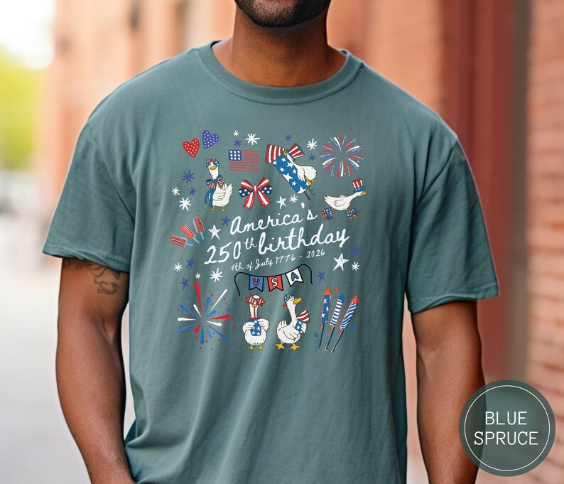 May include: A blue spruce t-shirt featuring a graphic commemorating America's 250th birthday. The design includes fireworks, patriotic geese, and the text "America's 250th birthday 4th of July 1776-2026" in red, white, and blue.