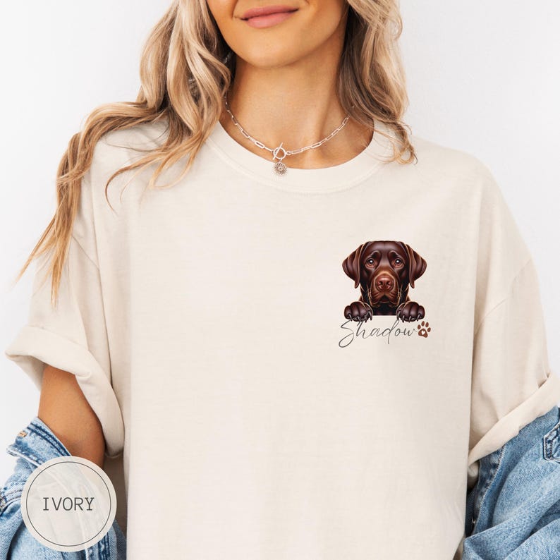 以下が含まれることがあります： 白いTシャツに、茶色のラブラドールレトリバーが上の方から顔を出しています。犬の名前「シャドウ」は、犬の下に黒い筆記体で書かれています。Tシャツの左下には「アイボリー」と表示されています。