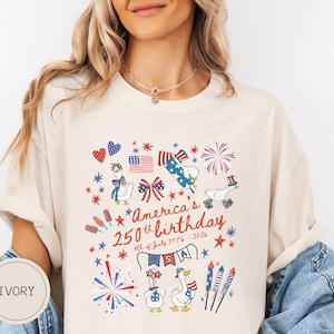 May include: Ivory t-shirt featuring a patriotic design for America's 250th birthday. The graphic includes fireworks, flags, and cartoon geese in red, white, and blue. Text reads "America's 250th birthday 4th of July 1776-2026".