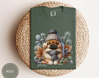 May include: A green t-shirt with a cute illustration of a Pekingese dog wearing a winter hat and scarf. The dog is surrounded by winter foliage and berries. The text "Sam" is printed below the dog.
