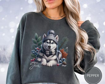 Puede incluir: Sudadera gris oscuro con un Husky siberiano que lleva una bufanda y un gorro a rayas grises y blancas. El perro está rodeado de follaje invernal y bayas. La palabra "Sam" está escrita en cursiva.