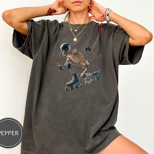 May include: Dark grey t-shirt featuring a skeleton roller skating with protective gear. The skeleton is wearing a helmet, knee pads, and roller skates. The word "Taylor" is written in cursive. The shirt has a Pepper label.