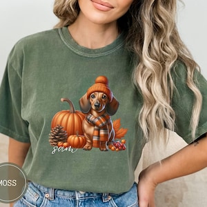 Op de afbeelding: Een groen T-shirt met een teckel die een bruin-oranje sjaal en een bruin-oranje hoed draagt. De teckel staat naast een pompoen en een dennenappel. De tekst "Sam" is op het T-shirt gedrukt.