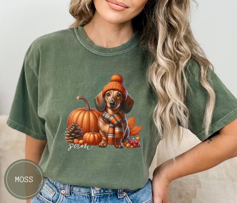 Op de afbeelding: Een groen T-shirt met een teckel die een bruin-oranje sjaal en een bruin-oranje hoed draagt. De teckel staat naast een pompoen en een dennenappel. De tekst "Sam" is op het T-shirt gedrukt.