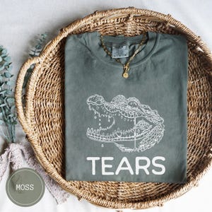 May include: A moss green short-sleeve t-shirt featuring a white line art graphic of an alligator with a single tear falling from its eye. The word TEARS is printed in large white capital letters below the alligator graphic on the front.