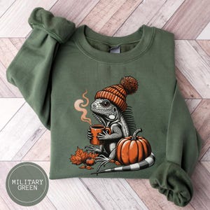 Könnte beinhalten: Ein grüner Pullover mit einem Aufdruck eines Leguans, der eine rot-weiße Strickmütze trägt und eine Tasse Kaffee hält. Der Leguan sitzt neben einem großen orangefarbenen Kürbis. Der Text "MILITARY GREEN" ist in der linken unteren Ecke des Bildes gedruckt.