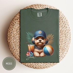 Può includere: Una maglietta verde con un'illustrazione di un cane cartone animato che indossa un cappello blu e tiene una palla da spiaggia. Il cane è circondato da piante tropicali e un bicchiere di succo d'arancia. Il testo "Sam" è scritto sotto il cane.