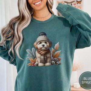 Op de afbeelding: Een tealgroen sweatshirt met een afbeelding van een witte hond die een bruin-witte gebreide muts en een bruine sjaal draagt. De hond is omringd door herfstbladeren en dennenappels.
