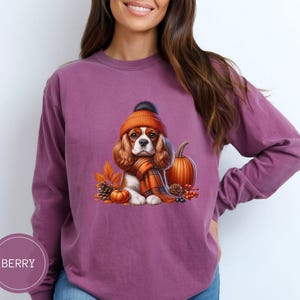 May include: A purple long-sleeved sweatshirt with a graphic of a Cavalier King Charles Spaniel wearing an orange knit hat and scarf, holding a small pumpkin. The dog is surrounded by fall foliage, including acorns, leaves, and berries.