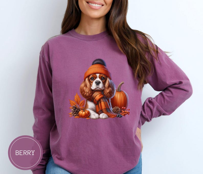 May include: A purple long-sleeved sweatshirt with a graphic of a Cavalier King Charles Spaniel wearing an orange knit hat and scarf, holding a small pumpkin. The dog is surrounded by fall foliage, including acorns, leaves, and berries.