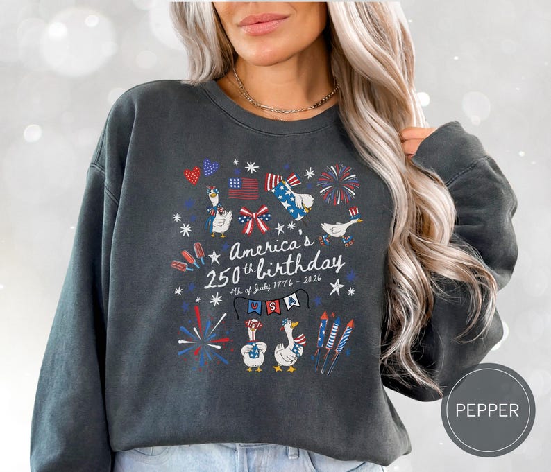 May include: Dark grey sweatshirt featuring a patriotic design for America's 250th birthday. The design includes fireworks, flags, and cartoon geese. Text reads "America's 250th birthday 4th of July 1776-2026".