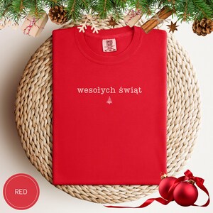 Może przedstawiać: Czerwony t-shirt z kr&oacute;tkim rękawem z białym napisem wesołych świąt oraz małą białą ikoną choinki wyśrodkowaną na klatce piersiowej. Koszulka posiada okrągły dekolt oraz metkę marki Comfort Colors nadrukowaną po wewnętrznej stronie kołnierza.