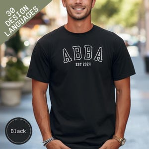Pode incluir: Camiseta preta de gola redonda com a palavra "ABBA" em branco, letras de bloco delineadas, e o texto "EST 2024" abaixo. A camiseta é de cor preta lisa. Uma etiqueta circular no canto inferior esquerdo diz "Black".