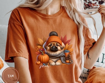 May include: Orange t-shirt with a cute cartoon pug wearing a brown and red knit hat and a gray scarf. The pug is surrounded by fall leaves, pumpkins, and acorns. The text "Sam" is printed below the pug.