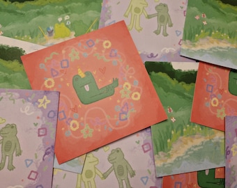 May include: A collection of colorful art prints featuring whimsical frog illustrations. The central print is coral-colored with a green frog wearing a unicorn horn. Other prints show frogs holding hands and a frog fishing. The background has a painted landscape.