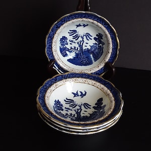 Op de afbeelding: Een stapel blauw-witte porseleinen kommen met een geschulpte rand en gouden rand. Elke kom heeft een landschapsontwerp met bomen, gebouwen en vogels. De kommen worden op een donkere ondergrond gepresenteerd.