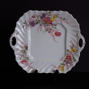 Puede incluir: Plato cuadrado blanco para servir con borde festoneado y diseño floral. El plato presenta flores coloridas en tonos rosa, morado, amarillo y rojo. Tiene dos asas y está sobre un soporte negro.