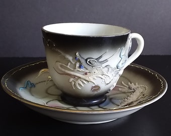May include: A white teacup and saucer set with a dragon design. The cup and saucer are white with a black gradient and gold trim. The dragon is detailed with blue, gray, and gold accents. The set is on a black surface.