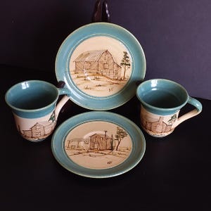 May include: A ceramic tea set with a rustic design. The set includes two mugs and two plates, each featuring a hand-drawn illustration of a barn or cabin. The rims are a teal color, and the illustrations are in shades of brown. The initials "WE" are visible.