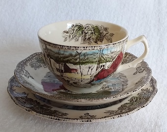 Può includere: Un set vintage di tazza da tè e piattino con una scena invernale che presenta un fienile rosso e alberi coperti di neve. Il set è decorato con un motivo floreale marrone e verde.
