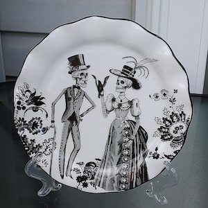 Può includere: Piatto in ceramica bianca con bordo smerlato nero, con un'illustrazione in bianco e nero di due scheletri in abito formale. Il piatto ha un design floreale ed è esposto su un supporto trasparente.