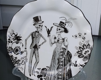 Puede incluir: Plato de cerámica blanco con borde festoneado negro, con una ilustración en blanco y negro de dos esqueletos con vestimenta formal. El plato tiene un diseño floral y se exhibe sobre un soporte transparente.