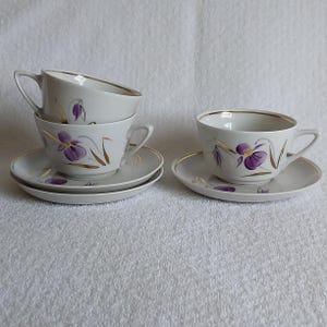 May include: A set of three white porcelain teacups and two matching saucers, each decorated with painted purple iris flowers and gold leaf accents. The cups feature curved handles and gold-trimmed rims, with one cup resting atop another.