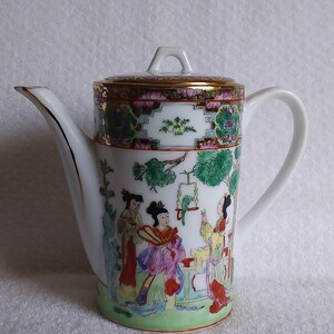 May include: A white porcelain teapot featuring a cylindrical body and a lid with a gold-trimmed rim. The exterior is decorated with a colorful hand-painted scene of three figures in traditional attire, a green bird in a cage, and foliage.