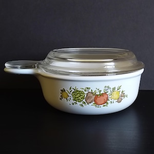 May include: A white ceramic casserole dish with a single handle and a clear glass lid. The exterior features a colorful botanical illustration of various vegetables including a tomato, artichoke, and lemon, arranged in a horizontal band.