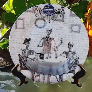 Op de afbeelding: Een decoratieve plaat met een skeletthema. De plaat toont drie skeletten die rond een tafel zitten, met de woorden "Bone Appetit" op een klein bordje. De plaat heeft een witte achtergrond met script en ingelijste afbeeldingen. Gemaakt in Engeland.