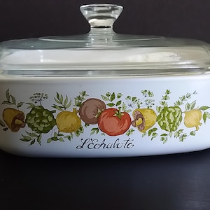 May include: A rectangular white ceramic casserole dish with a clear glass lid and a knob handle. The exterior features a colorful botanical illustration of various vegetables and herbs, with the text L'Echalote printed in black script lettering.