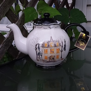 May include: White ceramic teapot with a black lid and handle. The teapot features a spooky haunted house design with bats and bare trees. The tag reads "Spooky Night".