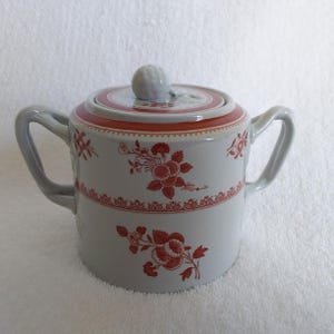 May include: A white ceramic sugar bowl with a lid, featuring red floral patterns and accents. The bowl has two handles and a knob on the lid. The design includes a red band around the top and bottom, with a delicate floral motif.