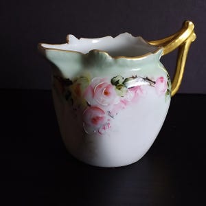 May include: A white porcelain creamer with a gold handle and rim. The creamer features a hand-painted design of pink roses and green leaves on a pale green background.