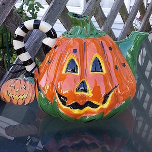 May include: A ceramic Halloween teapot in the shape of a jack-o'-lantern. The orange pumpkin-shaped body has a smiling face with black eyes and a black moustache. The handle is black and white striped, and the spout is green.