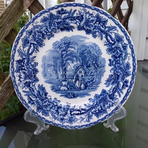 May include: A round ceramic plate featuring a blue and white transferware pattern. The center depicts a rural landscape with a cottage, trees, and figures. The wide rim is decorated with a detailed border of grapevines and leaves.