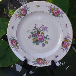 Può includere: Un piatto di porcellana bianca con bordo dorato e motivo floreale. Il piatto presenta un bouquet centrale di rose rosa, tulipani viola e altri fiori colorati. Ulteriori gruppi floreali sono disposti sul bordo.