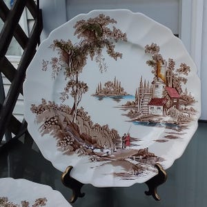 May include: A white ceramic dinner plate featuring a brown and blue landscape scene. The scene depicts a small cottage with a red roof, a river, and trees. The plate has a scalloped edge.