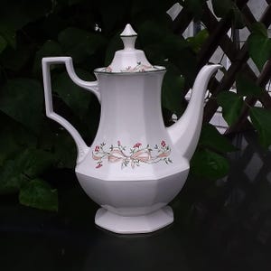 May include: A white porcelain coffee pot with a faceted, octagonal body and a matching lid. The surface features a delicate floral pattern with pink flowers, green leaves, and a light pink ribbon motif. It has a tall handle and a curved spout.